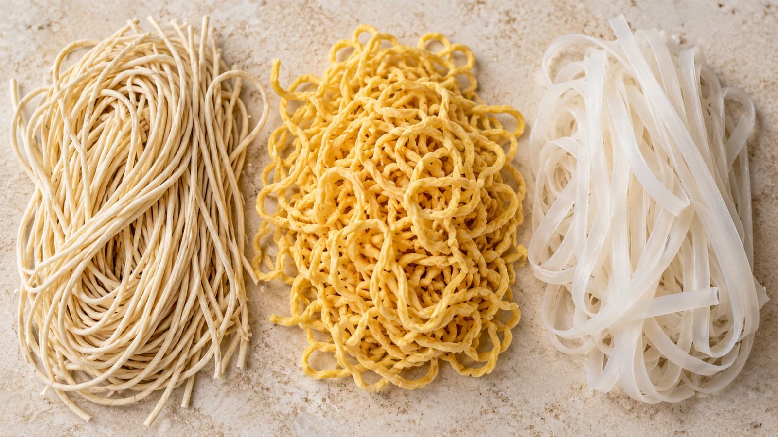 Three separate portions of dried wheat noodles, yellow egg noodles, and flat rice noodles showing differences in shape and color.